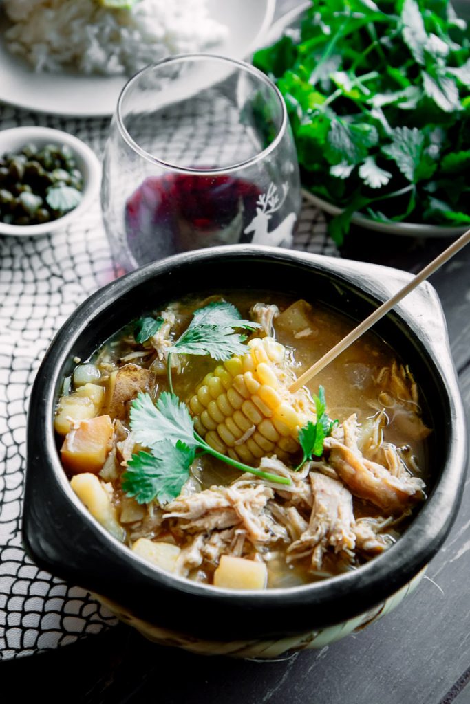 A chamba pot with Colombian ajiaco on a wood table with a side of rice, avocado, cream, and cilantro and a glass of wine.