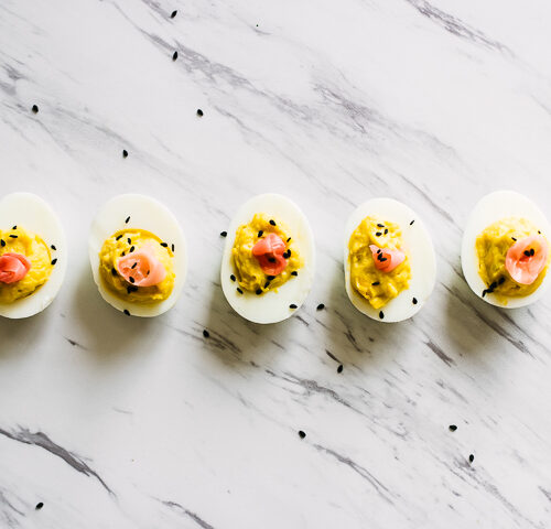 A wasabi ginger deviled eggs with pickled ginger on top on a slab of marble garnished with black sesame seeds.
