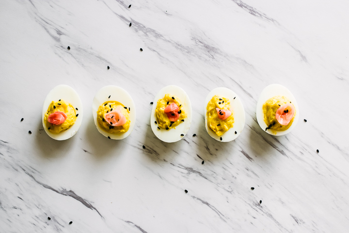 A wasabi ginger deviled eggs with pickled ginger on top on a slab of marble garnished with black sesame seeds.