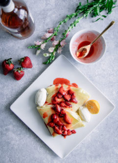 A crepe with ricotta and vanilla bourbon strawberries on a white plate on a blue table with flowers.
