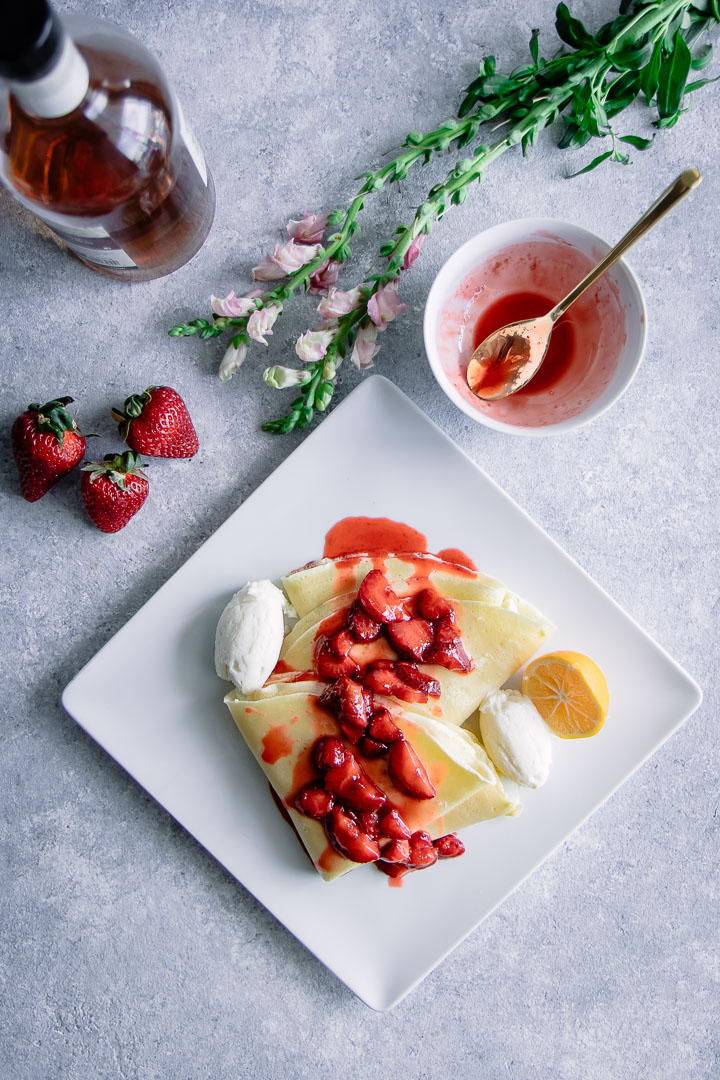 A crepe with ricotta and vanilla bourbon strawberries on a white plate on a blue table with flowers.