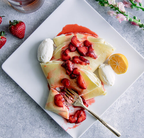 A crepe with ricotta and vanilla bourbon strawberries on a white plate on a blue table with flowers.