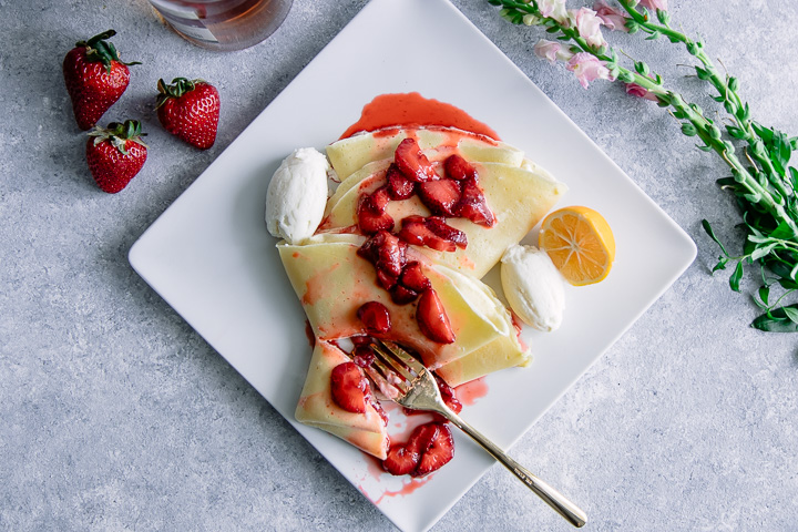 A crepe with ricotta and vanilla bourbon strawberries on a white plate on a blue table with flowers.