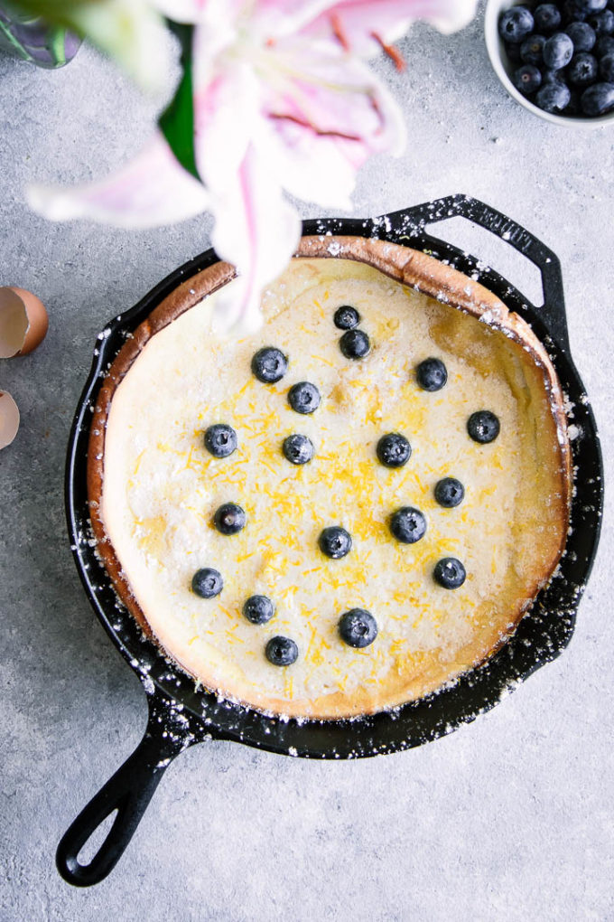 Dutch baby pancake with blueberries in a cast iron skillet on a blue table with pink flowers.