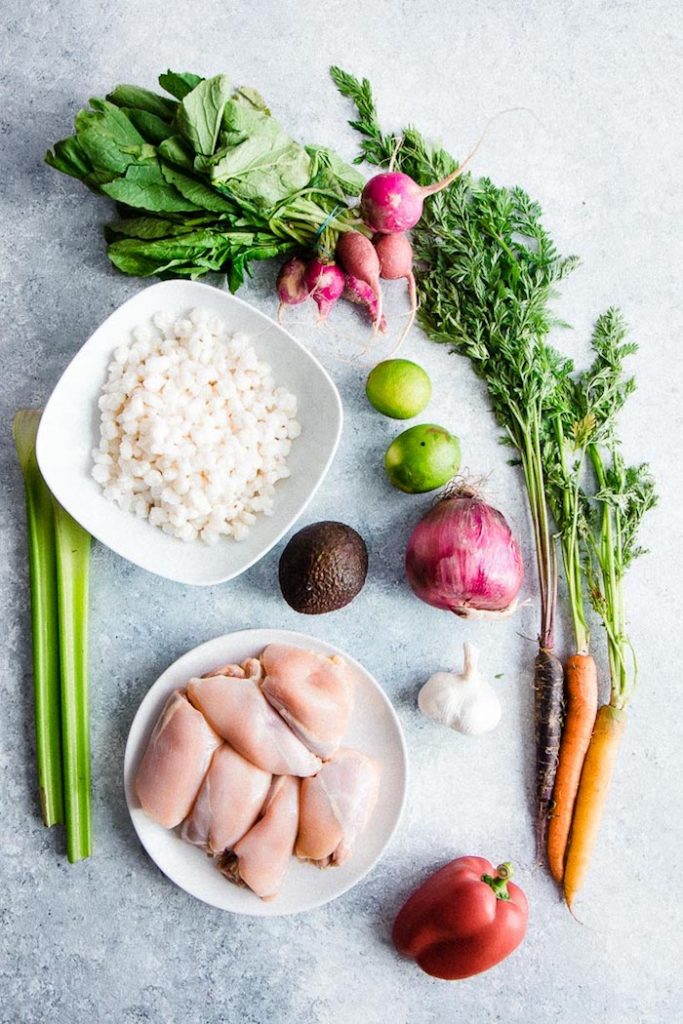 Chicken, hominy, celery, avocado, onion, garlic, limes, radishes and carrots on a table to make pozole verde.