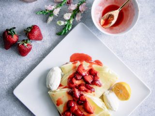 A square plate with a crepe with strawberry sauce on top with a bowl and a gold spoon and red flowers, with the words "ricotta crepe with vanilla bourbon strawberries" in black writing