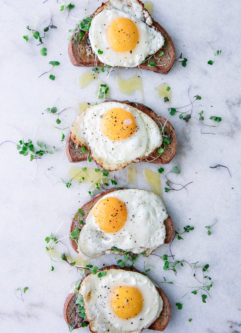 Four pieces of toast with olive tapenade, olive oil, microgreens, and a sunny egg on top of marble.