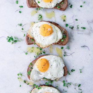Four pieces of toast with olive tapenade, olive oil, microgreens, and a sunny egg on top of marble.