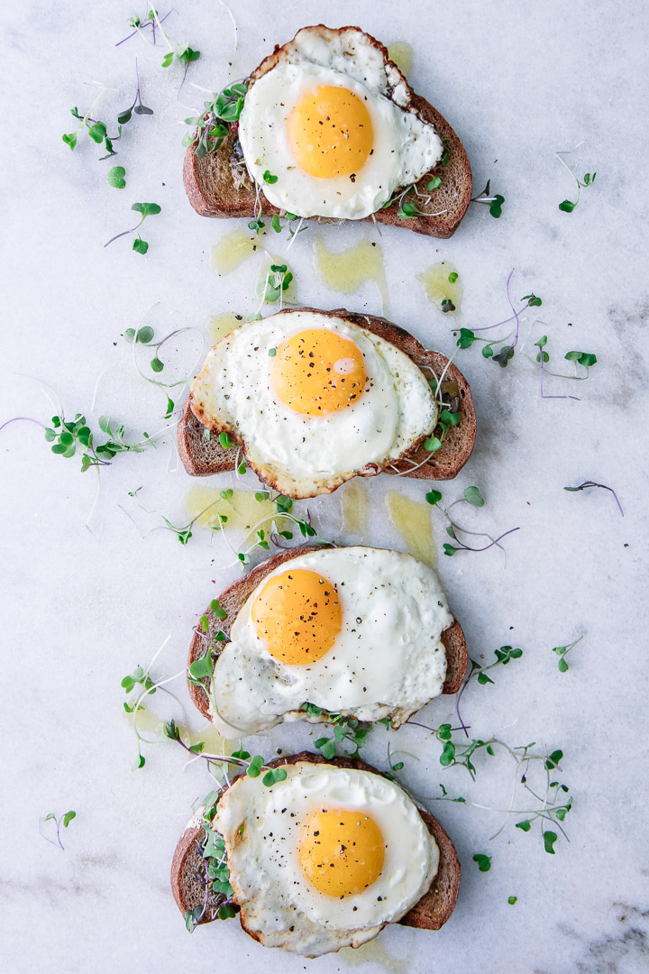 Four pieces of toast with olive tapenade, olive oil, microgreens, and a sunny egg on top of marble.