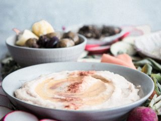 A bowl of white bean hummus dip with assorted vegetables and olives on a platter.