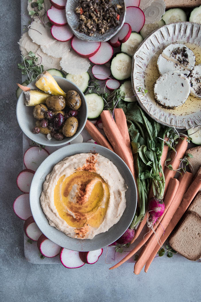 A mezze platter with white bean hummus, mixed olives, tapenade, raw vegetables, and assorted crackers on a blue table.