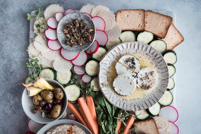 A mezze platter with white bean hummus, mixed olives, tapenade, raw vegetables, and assorted crackers on a blue table.