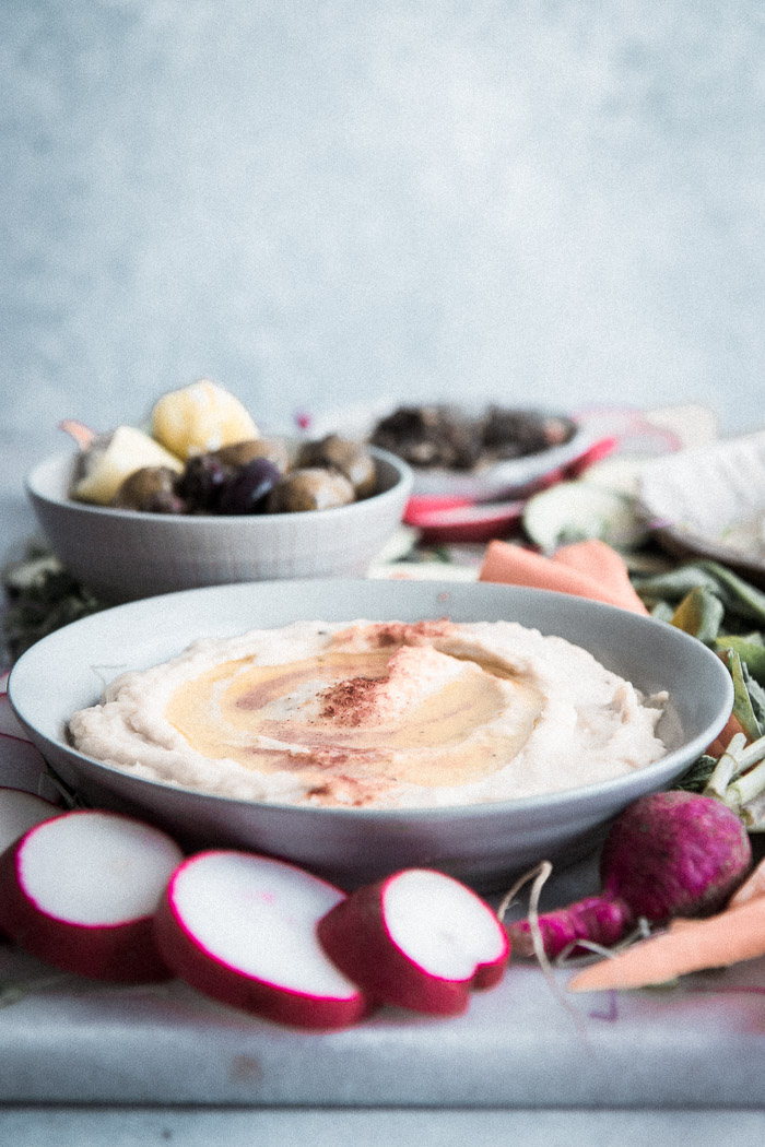 A bowl of white bean hummus dip with assorted vegetables and olives on a platter.