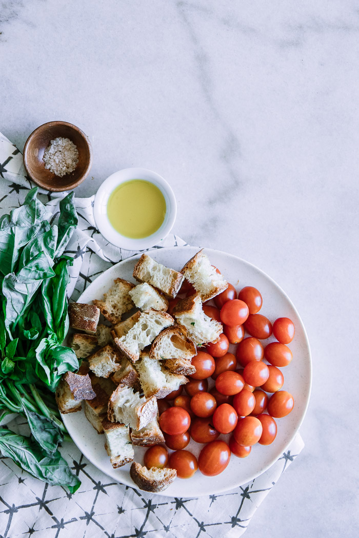 Cherry tomatoes, bread, basil, and olive oil for a caprese panzanella salad on a white table.