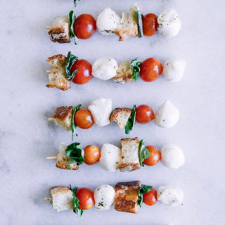Skewers with cherry tomatoes, mozzarella, basil, and bread crumbs on a stick on a white table.