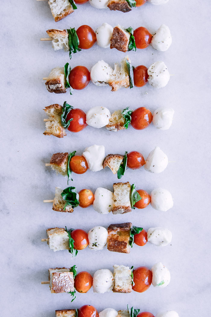 Skewers with cherry tomatoes, mozzarella, basil, and bread crumbs on a stick on a white table.