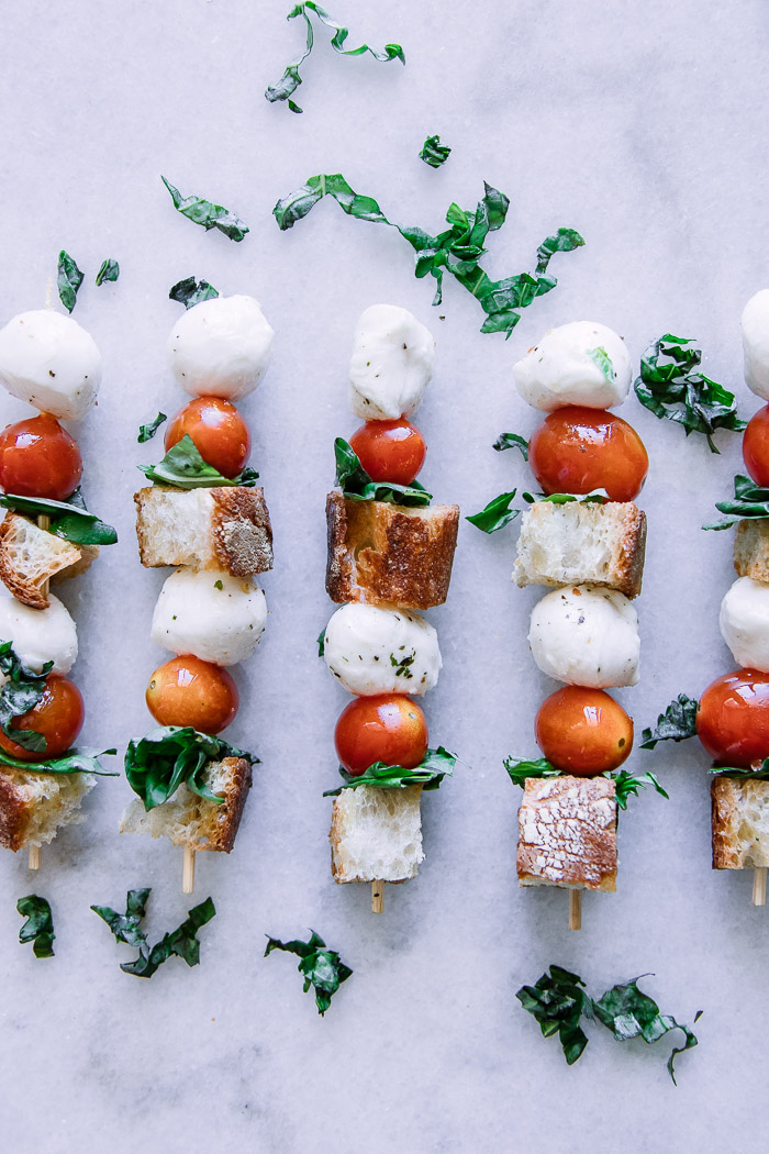 Skewers with cherry tomatoes, mozzarella, basil, and bread crumbs on a stick on a white table.