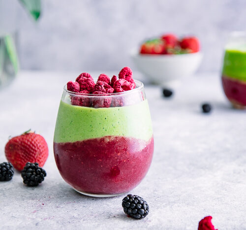 A green smoothie with avocado, spinach, banana, and raspberries on a white table with berries.