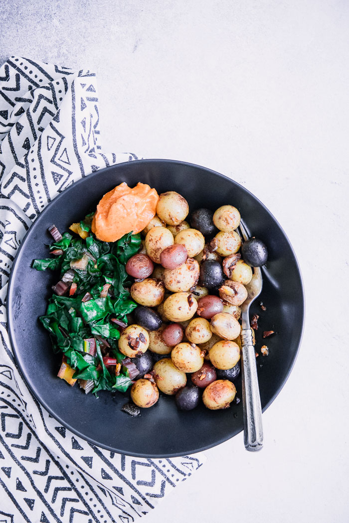 A black bowl with breakfast potatoes, cooked greens, and garlic aioli.