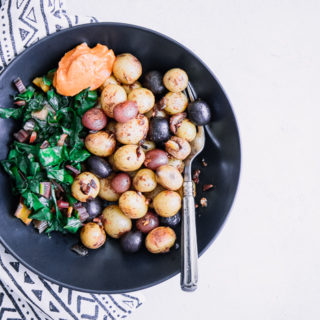 A black bowl with breakfast potatoes, cooked greens, and garlic aioli.