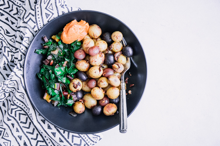 A black bowl with breakfast potatoes, cooked greens, and garlic aioli.