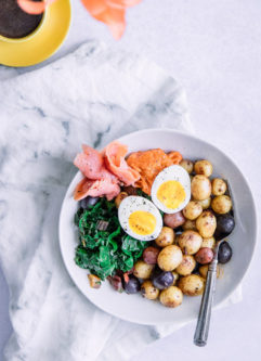 A breakfast bowl with salmon, potatoes, eggs, and chard in a white bowl on a white table with a cup of espresso.