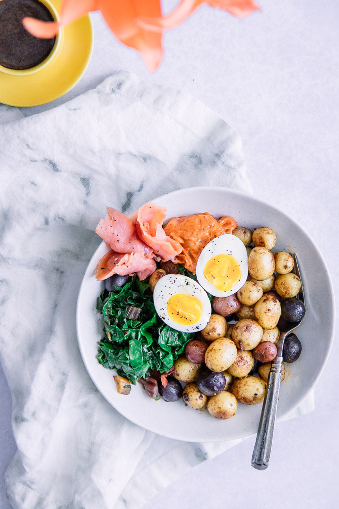 A breakfast bowl with salmon, potatoes, eggs, and chard in a white bowl on a white table with a cup of espresso.