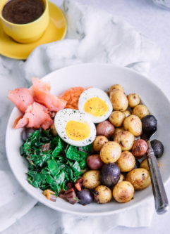 A brunch bowl with salmon, potatoes, greens, aioli, and egg on a white table with a yellow espresso cup.