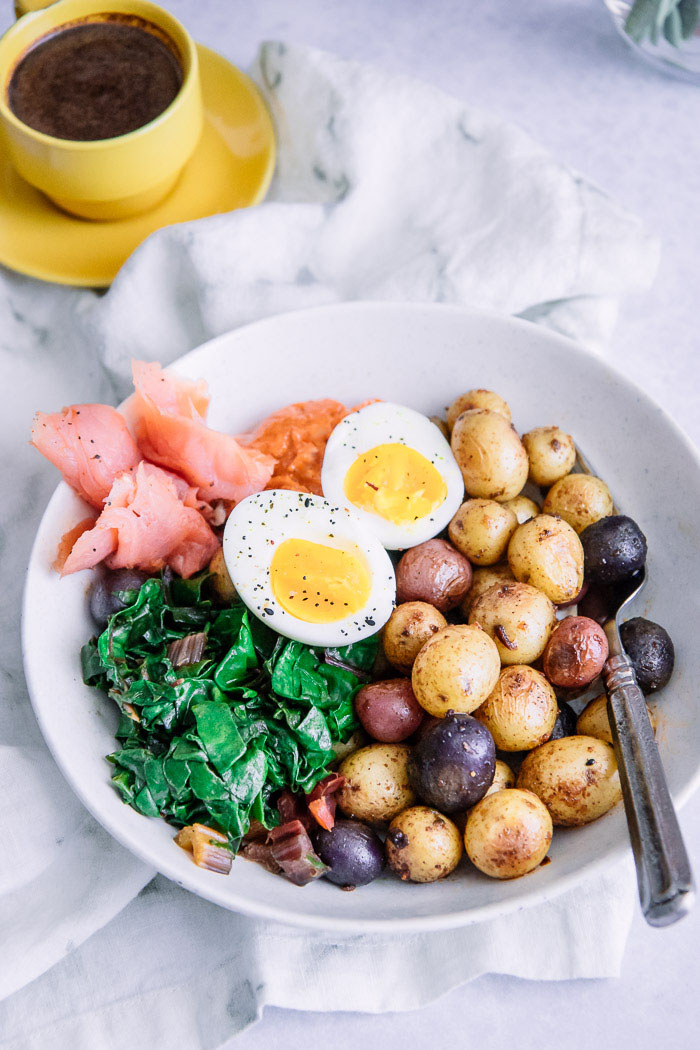 A brunch bowl with salmon, potatoes, greens, aioli, and egg on a white table with a yellow espresso cup.