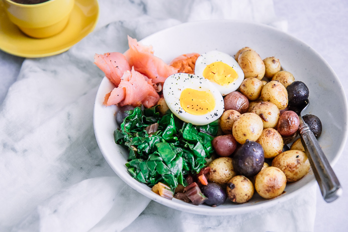 a breakfast for dinner bowl with salmon, eggs, and potatoes on a white table.