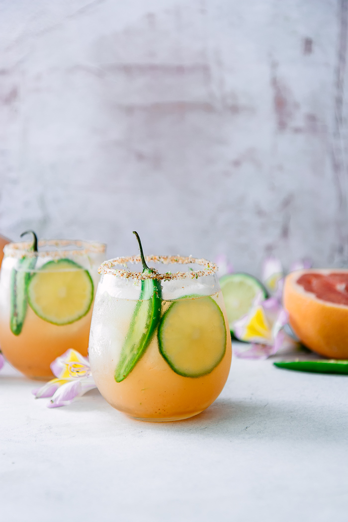 A margarita with grapefruit, lime, and orange with spicy jalapenos on a white table with flowers.