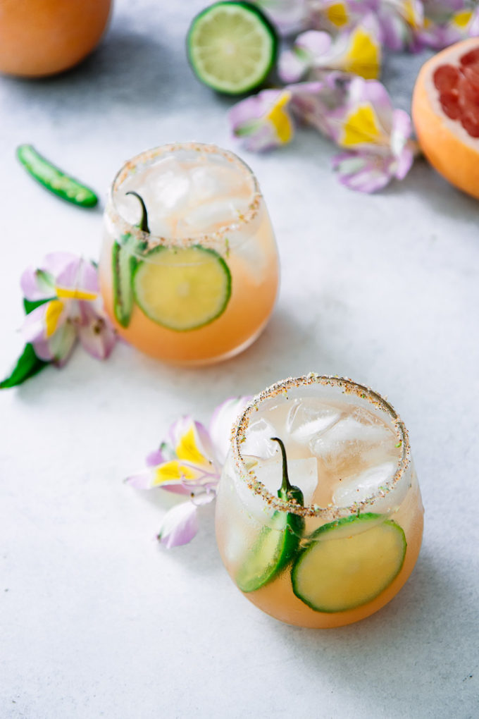 A margarita with grapefruit, lime, and orange with spicy jalapenos on a white table with flowers.