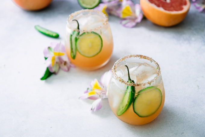 A margarita with grapefruit, lime, and orange with spicy jalapenos on a white table with flowers.