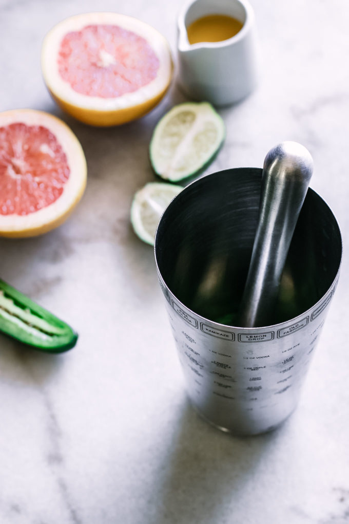 a cocktail shaker and muddler on a white table with grapefruits and limes