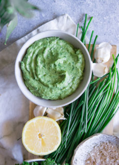 A bright green dip in a white bowl with a lemon and chives on a blue table.