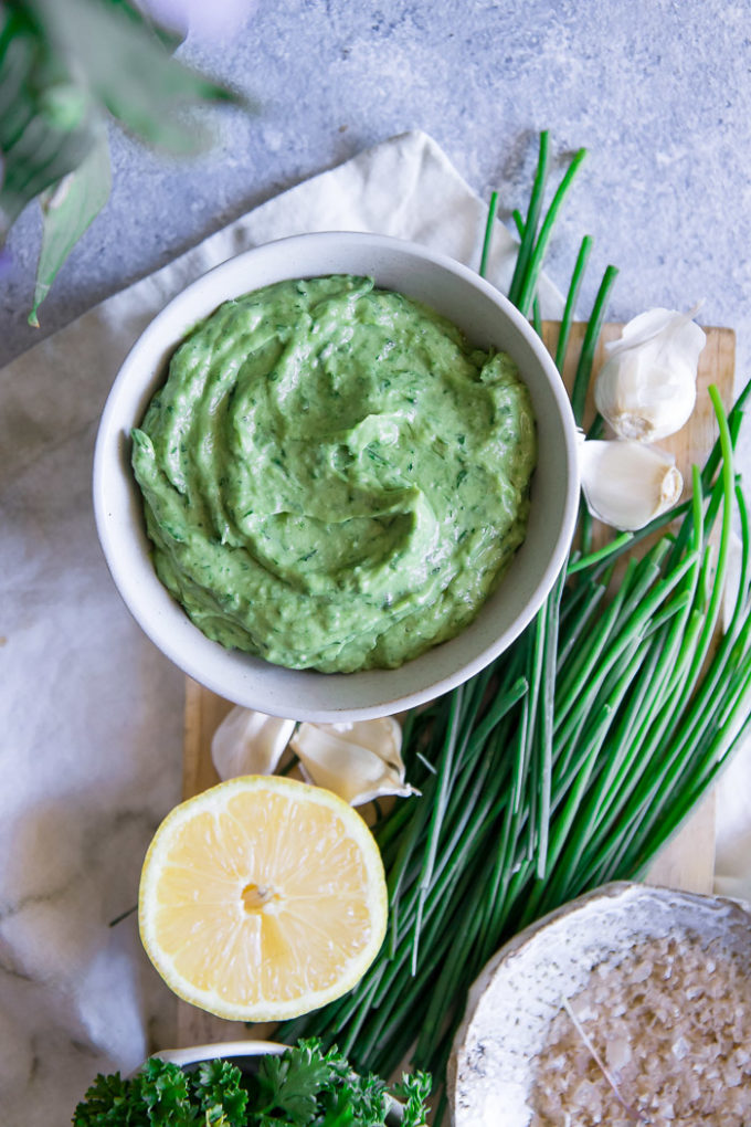 A bright green dip in a white bowl with a lemon and chives on a blue table.
