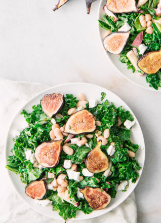 Two white salad plates on a white table with kale, figs, goat cheese, and white beans in a maple dressing.