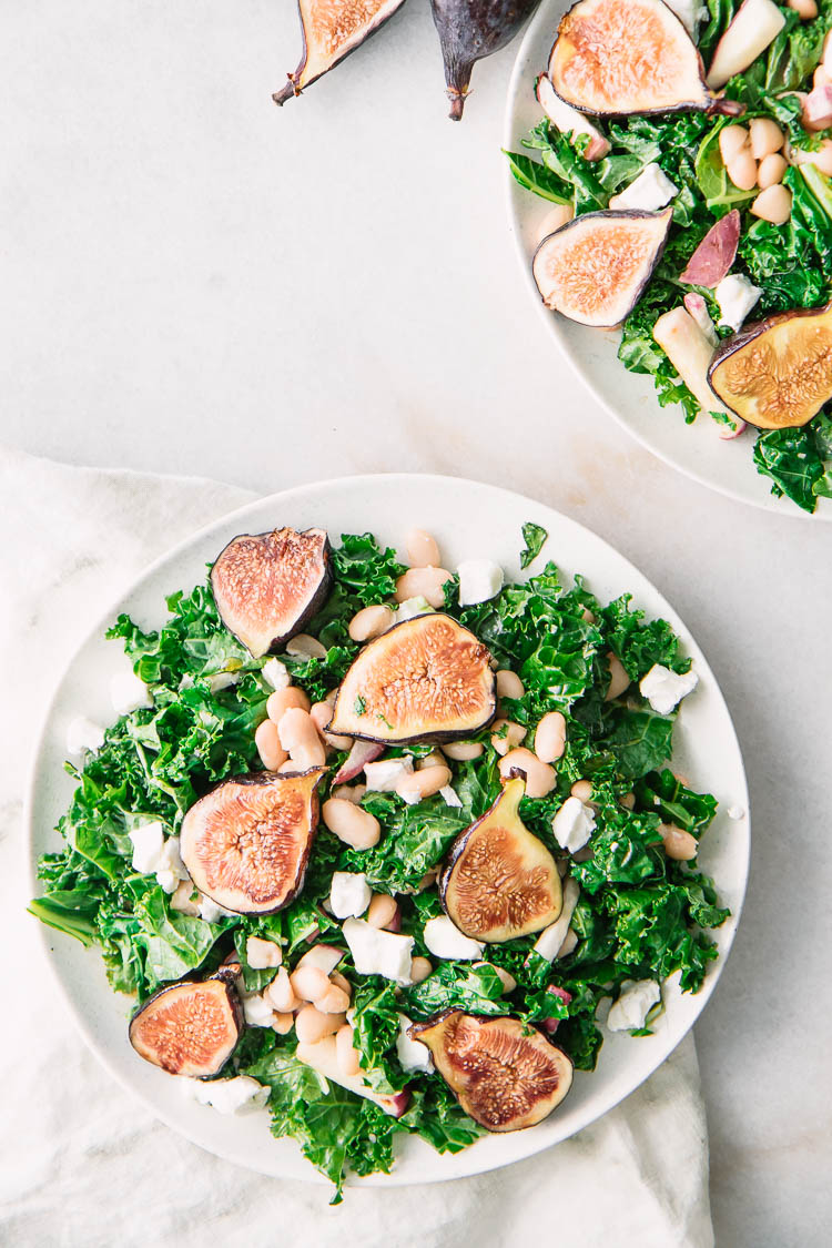 Two white salad plates on a white table with kale, figs, goat cheese, and white beans in a maple dressing.