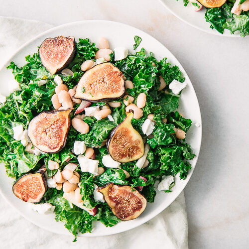 A green salad with kale, figs, white beans, and goat cheese on a white plate.