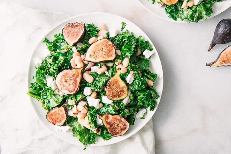 A green salad with kale, figs, white beans, and goat cheese on a white plate.
