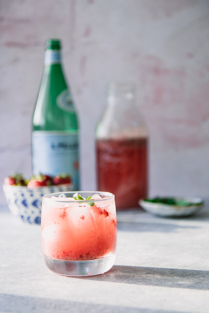 A strawberry and mint sparkling soda on a blue table with a bowl of strawberries and a bottle of sparkling water in the background.