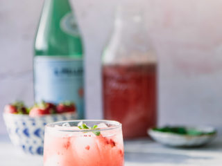 A sparkling water with strawberries and mint on a blue table with a pink background and the world "Strawberry Mint Spritzer" in black writing.