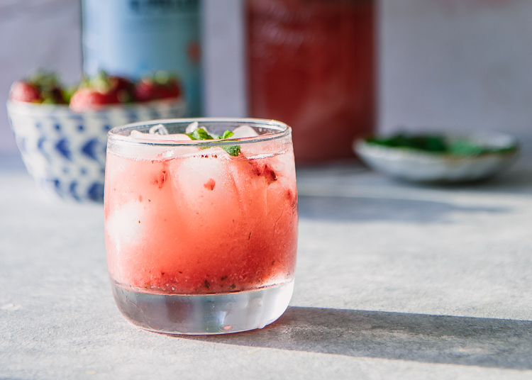 A glass of sparkling water with strawberries and mint on a blue table.