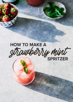 a cocktail glass filled with sparkling water, crushed strawberries, and mint on a blue table with sunlight beaming and a bowl of strawberries and a plate of mint leaves