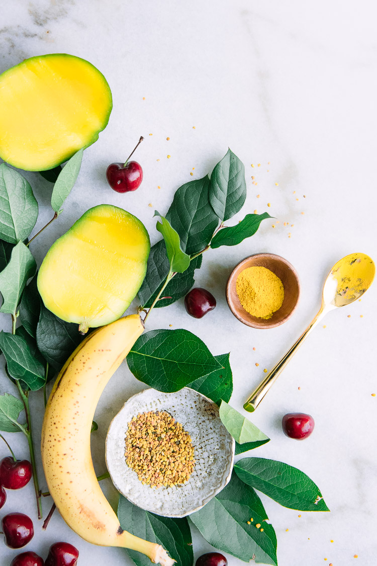 A flatlay of yellow superfoods including banana, mango, turmeric, bee pollen.