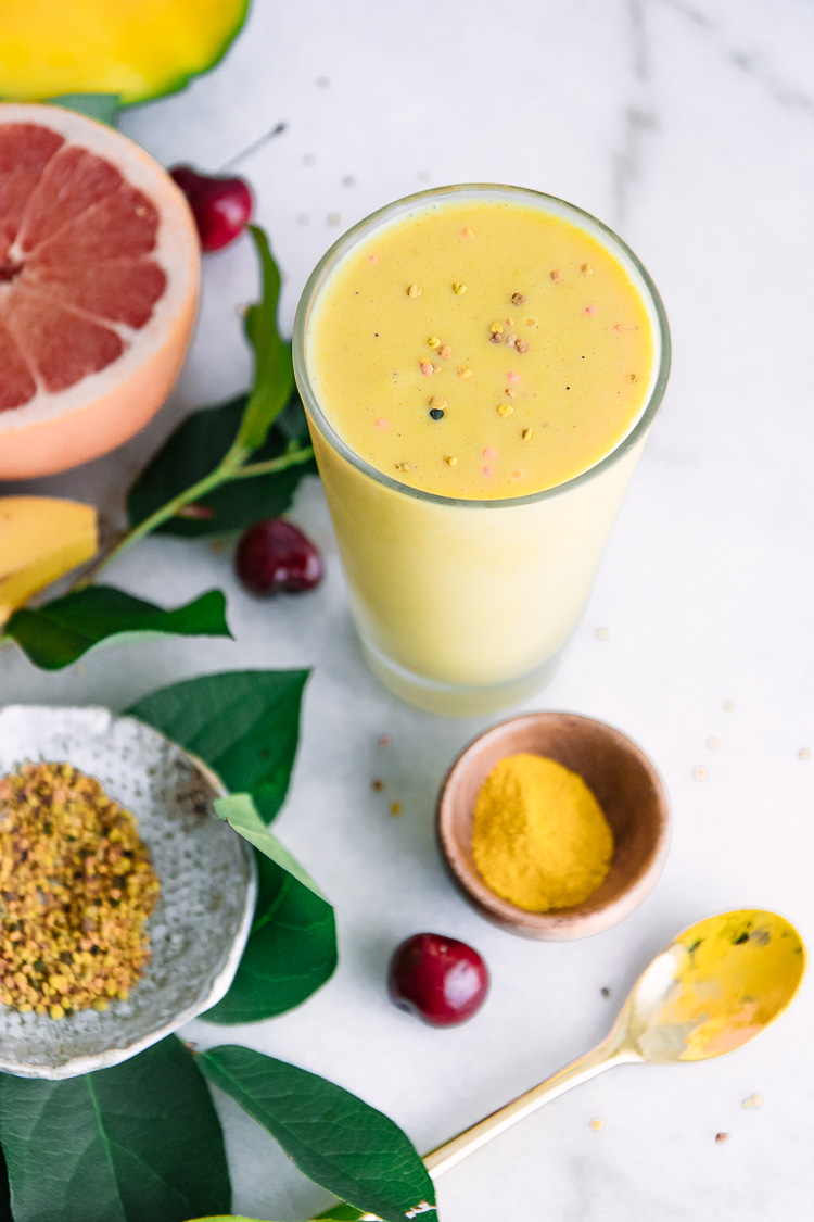 A mango banana pineapple yellow smoothie in a glass with bee pollen, turmeric, grapefruit, and bananas arrange around it on a white table.