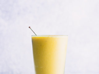 A ninety degree photo of a yellow mango banana smoothie in a glass with a white background.
