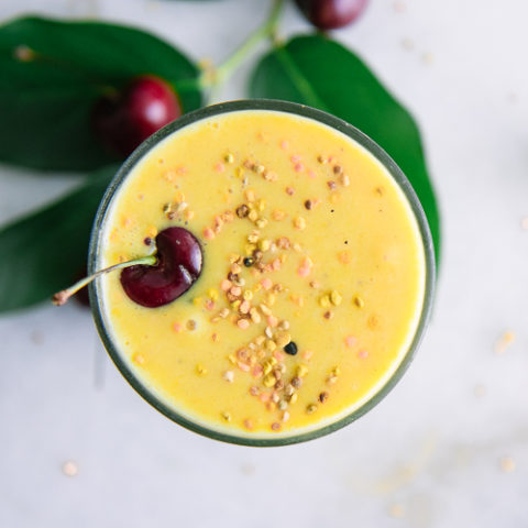 A top-down photo of a yellow smoothie in a glass with a sprinkle of bee pollen and a cherry as a garnish.