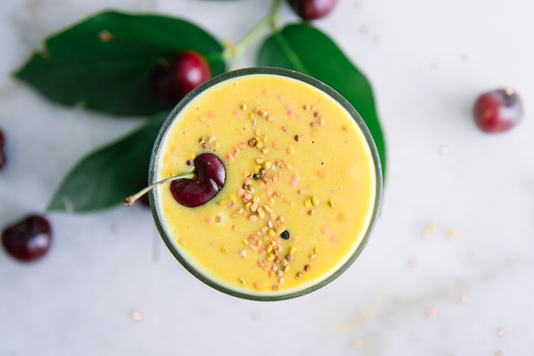 A top-down photo of a yellow smoothie in a glass with a sprinkle of bee pollen and a cherry as a garnish.