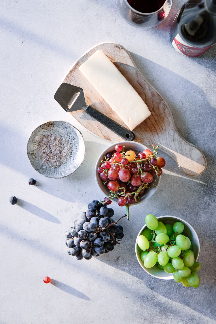Bowls of red, green, and purple grapes with parmesan cheese on a cutting board and a dish of pink smoky salt.